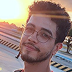 Hugo Vergnano é um homem branco, loiro, que usa bigode e tem cabelos lisos e castanhos. Ele usa uma camiseta preta e um pingente preto. Ele esta sorrindo e usando uma camiseta preta.