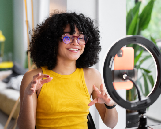 Uma
                pessoa com cabelo cacheado, usando uma blusa amarela, sorri
                e gesticula enquanto fala em frente a uma luz de anel. O
                ambiente parece ser interno, com iluminação suave e plantas
                ao fundo, sugerindo um espaço confortável, possivelmente
                para gravação de vídeos ou transmissões ao vivo.