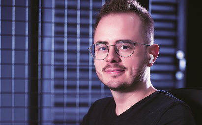 Lucas Colette, homem branco, de barba e cabelo curto loiros, tem olhos azuis e usa óculos de grau com o metal preto. Na foto, está sorrindo sem mostrar os dentes.
